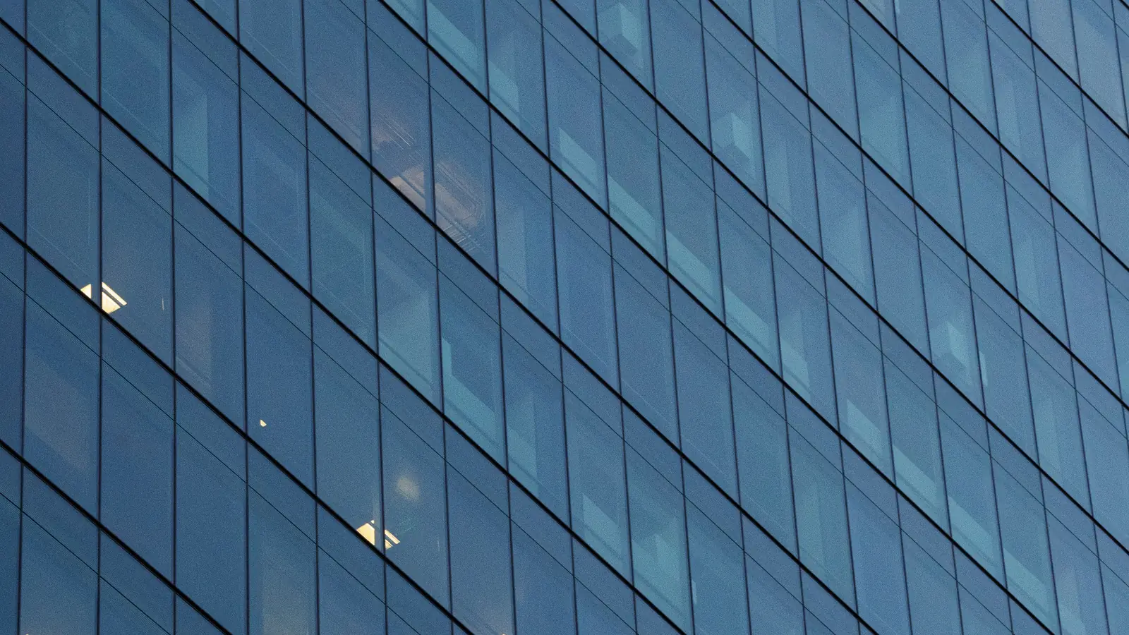 Glazen gevel van een modern kantoorgebouw met blauwe panelen en hier en daar licht achter de ramen